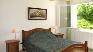 A bedroom with a wooden bed, two bedside tables with lamps, a green patterned bedspread, a framed floral painting on the wall, and a view of greenery through a window.