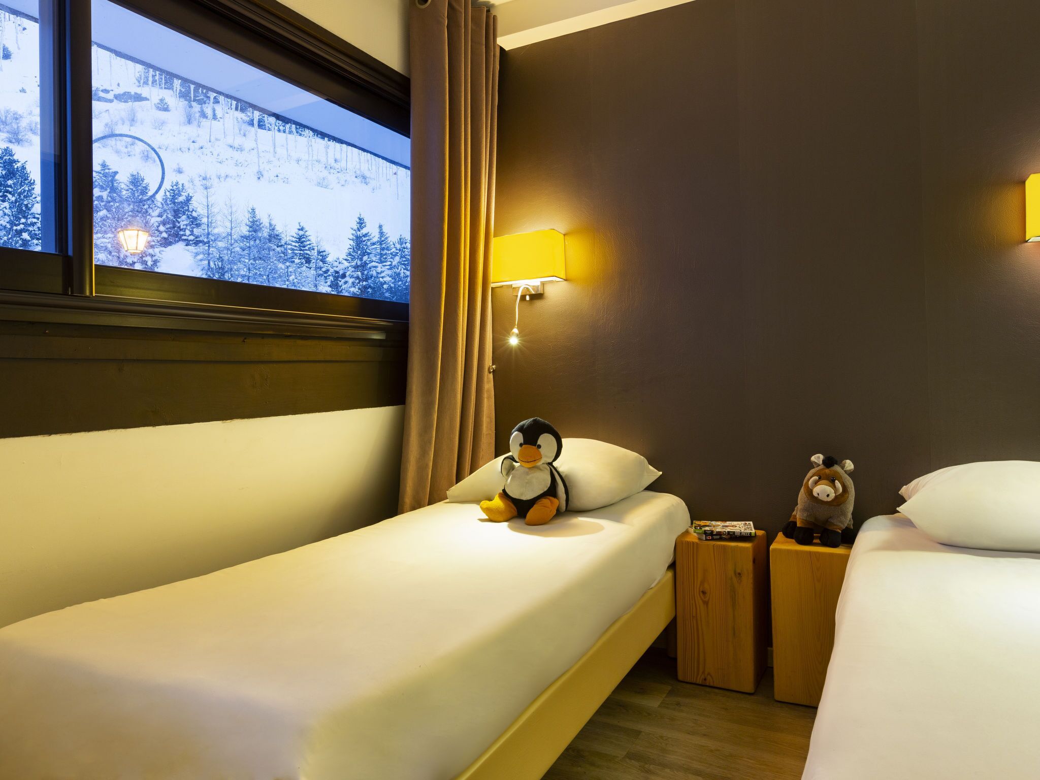 Ein Hotelzimmer mit zwei Betten, einem Fenster mit Blick auf eine verschneite Landschaft, einer Wandlampe und einem Stoffpinguin auf einem Bett.