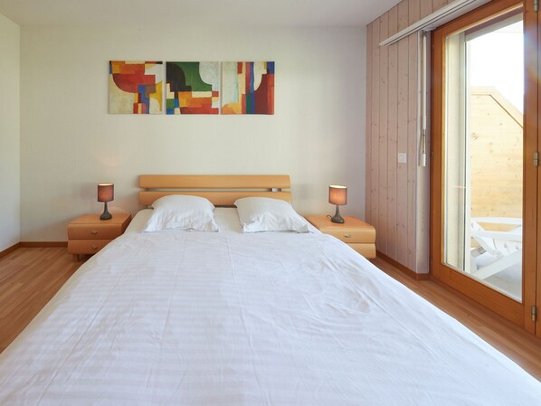 A bedroom with a large bed, two bedside tables with lamps, a wooden headboard, and a large window with a view of a wooden deck.