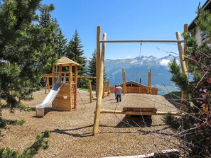 A playground with a slide and swings, surrounded by trees and a mountain view.