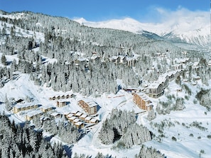 Un complejo turístico en la montaña, cubierto de nieve, con varias construcciones tipo chalet, rodeado de bosques frondosos y laderas nevadas.