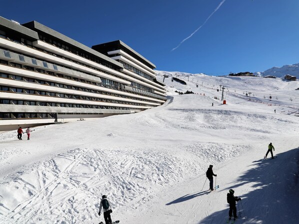 Ein Skigebiet mit einem modernen Gebäude, mehreren Balkonen und einer Skipiste mit Skifahrern.