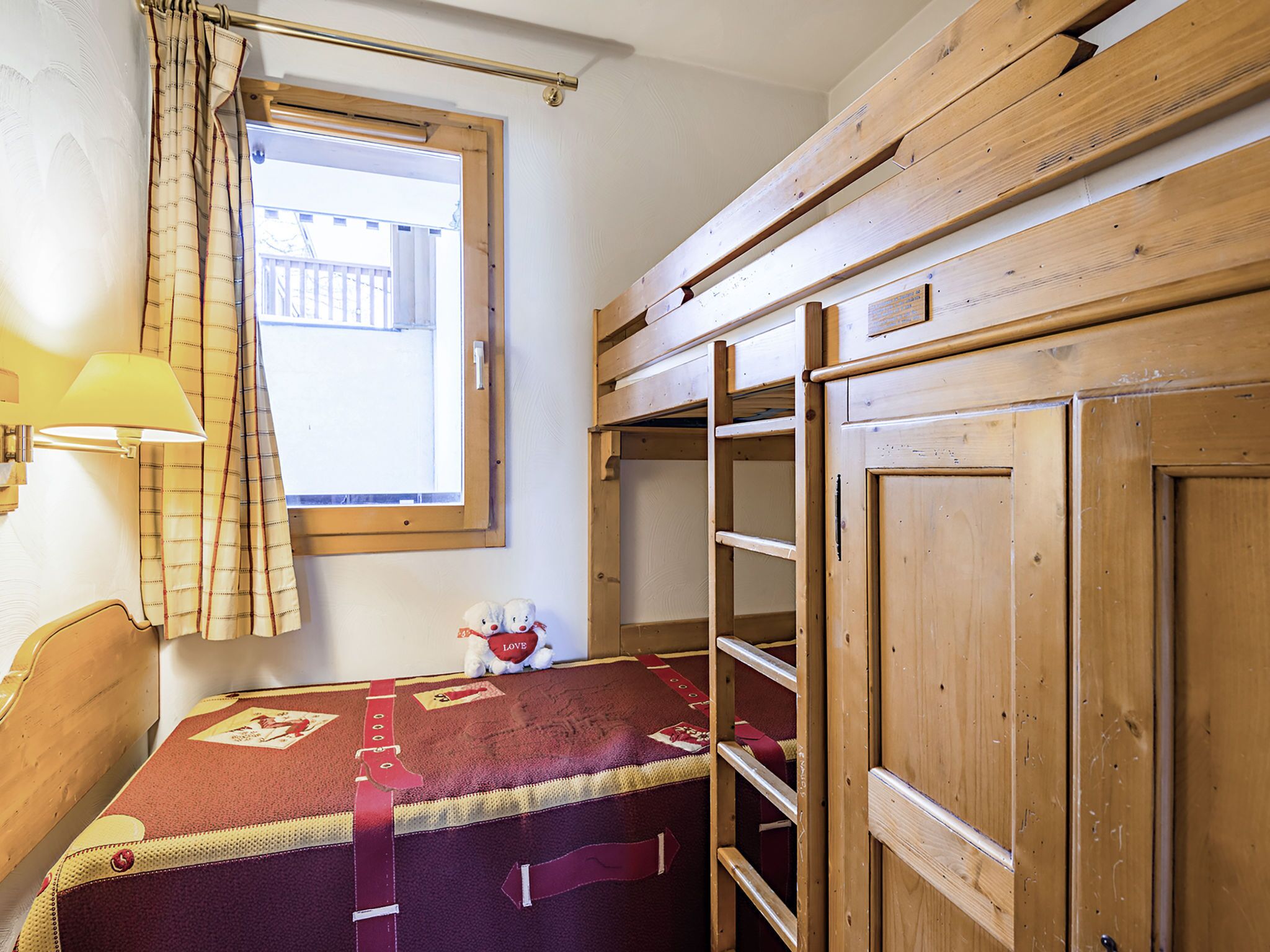 A room with a bunk bed, a window with curtains, a lamp, and a teddy bear on the bed.