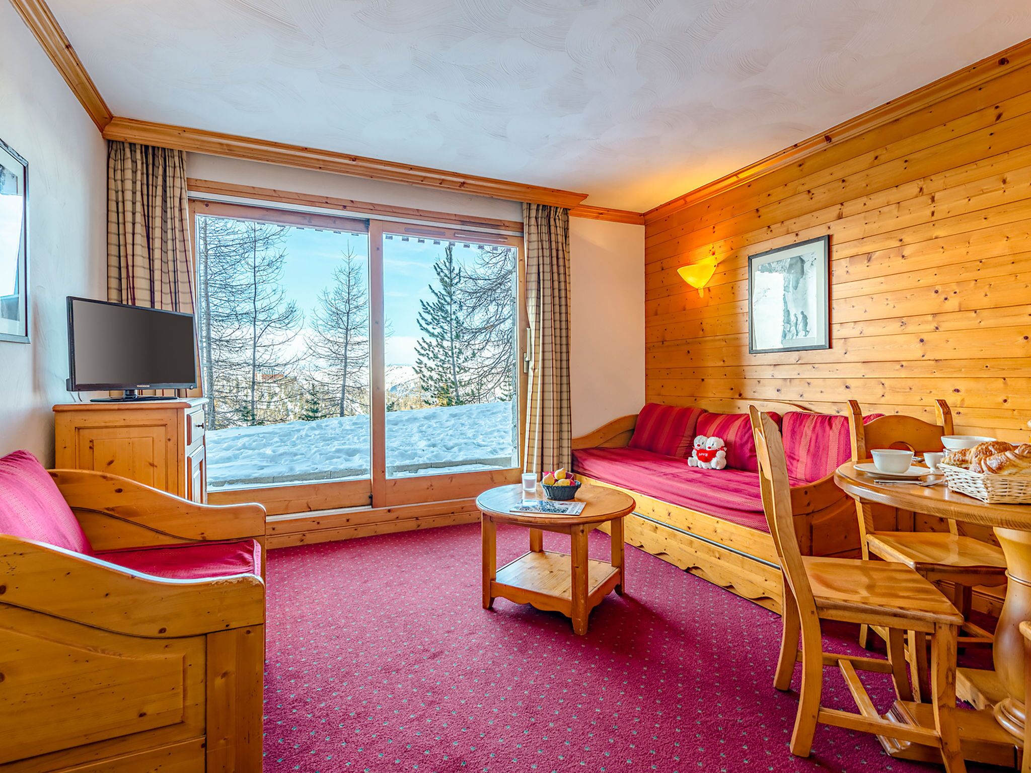 A cozy room with a wooden interior, a large window with a view of snow-covered trees, a television on a wooden cabinet, a small table with a bowl of fruit, and a red sofa with a teddy bear.