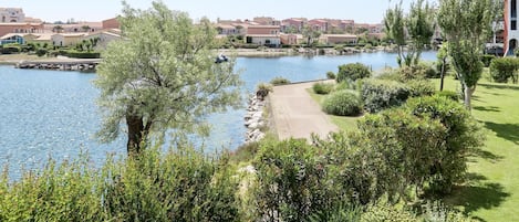 A lakeside view with a pathway leading to a dock, surrounded by lush greenery and residential buildings in the background.