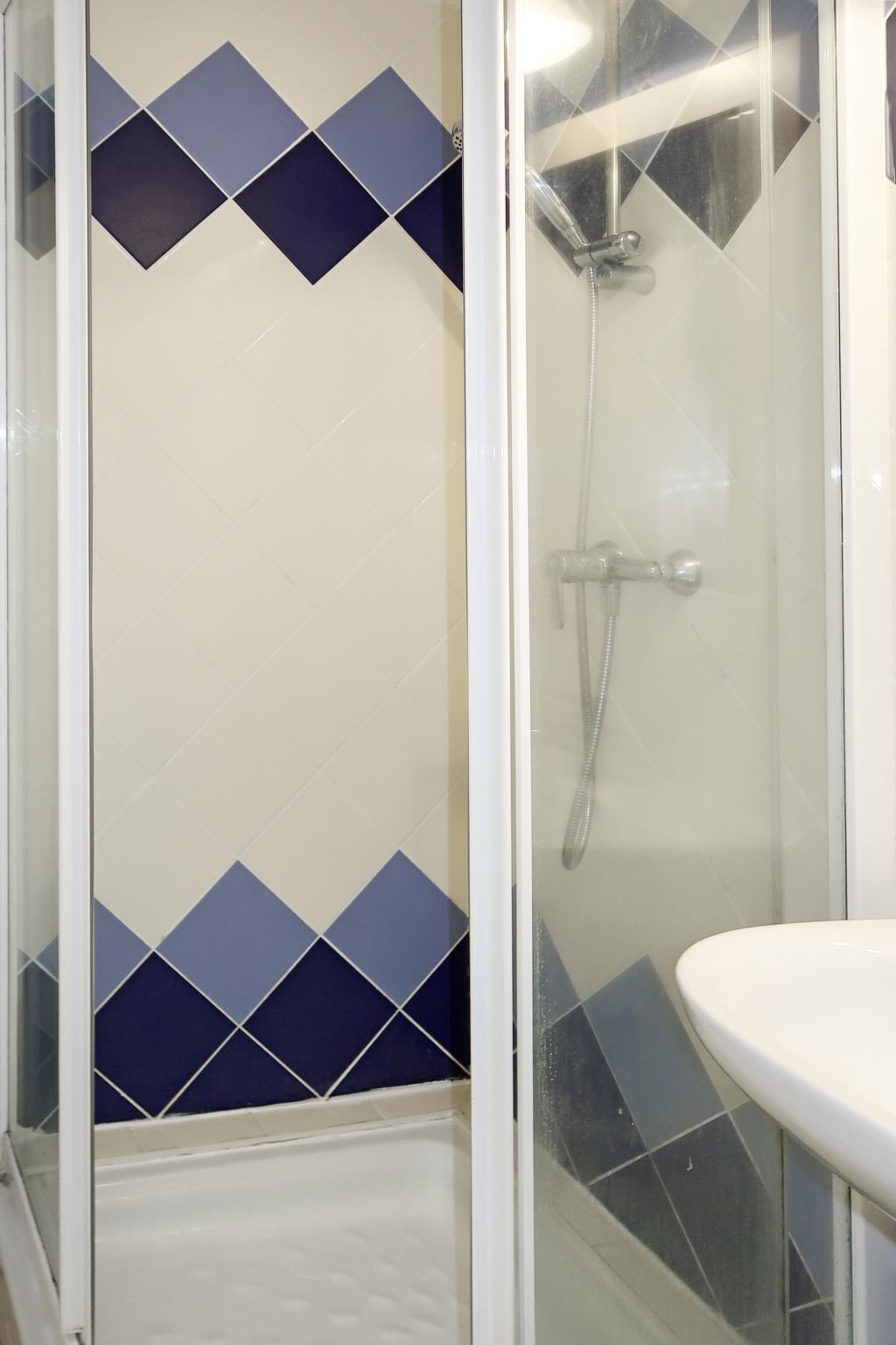 Un espace douche doté d’une porte en verre, d’un lavabo blanc et de carreaux bleus à motifs losangés sur les murs.