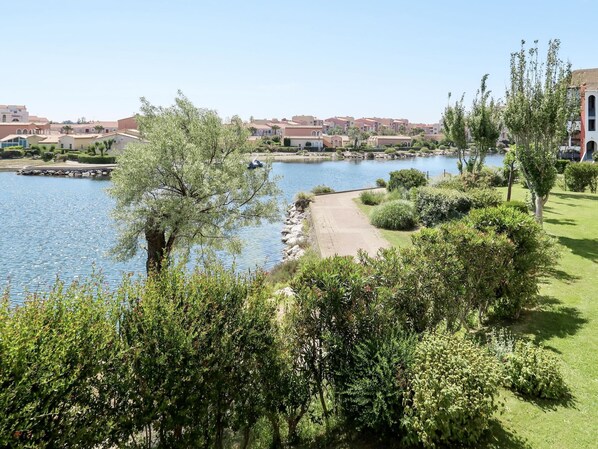 A lakeside view with a pathway leading to a dock, surrounded by lush greenery and residential buildings in the background.