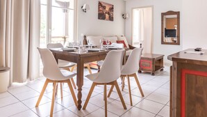 A dining area with a wooden table set for four, white chairs, and a sofa in the background. - Odalys Référence Le Domaine des Rois by Interhome (Bergheim)