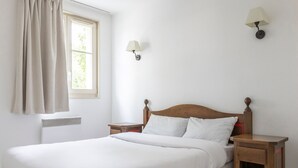 A neatly made bed with white linens in a room with white walls, a window with curtains, two bedside tables with lamps, and a radiator. - Odalys Référence Le Domaine des Rois by Interhome (Bergheim)