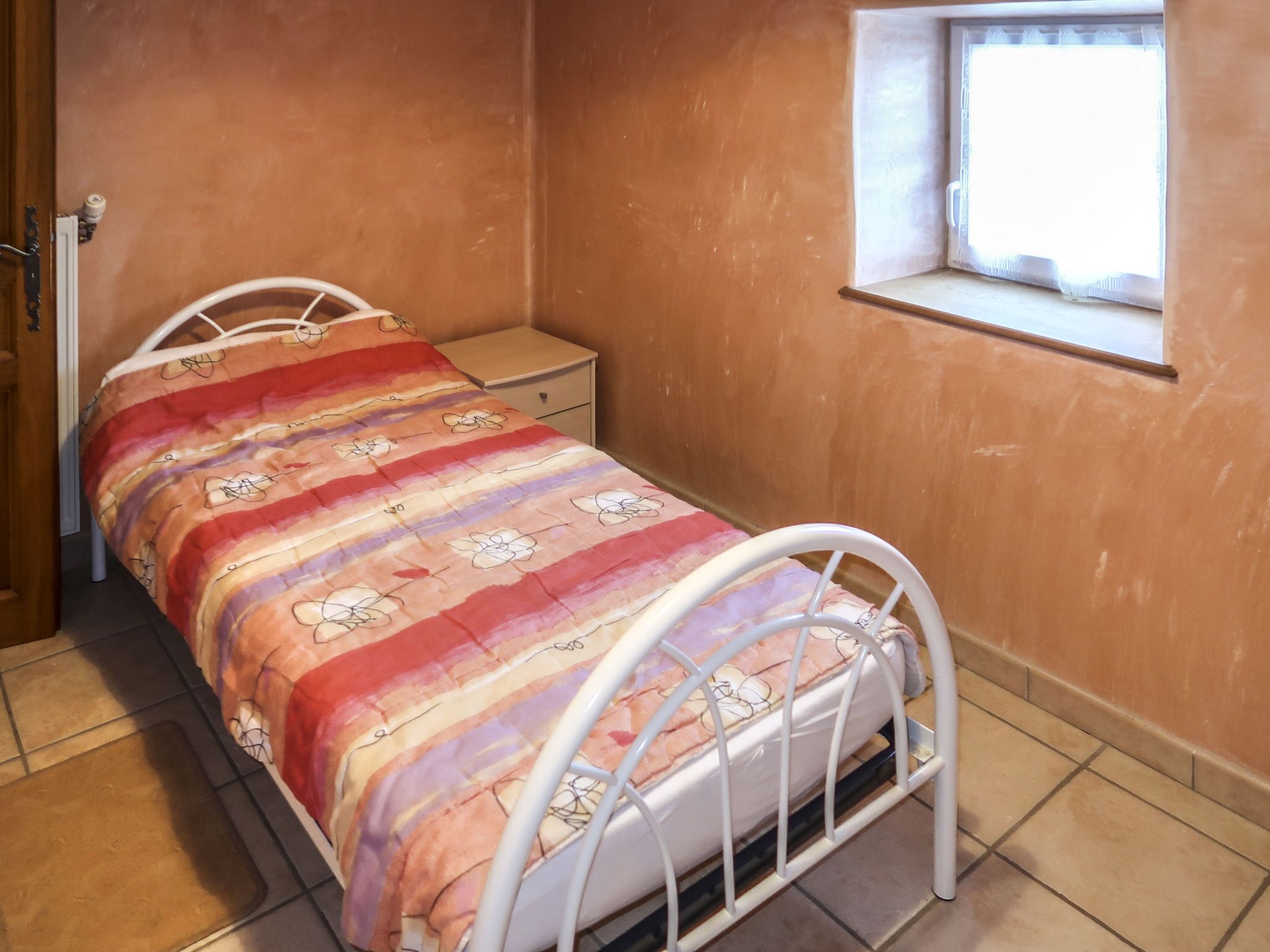 Una camera con un letto singolo, una struttura in metallo, un comodino, una finestra con tenda e una porta.