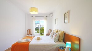 Een slaapkamer met een bed, nachtkastje, lamp, raam met gordijnen en uitzicht op gebouwen.