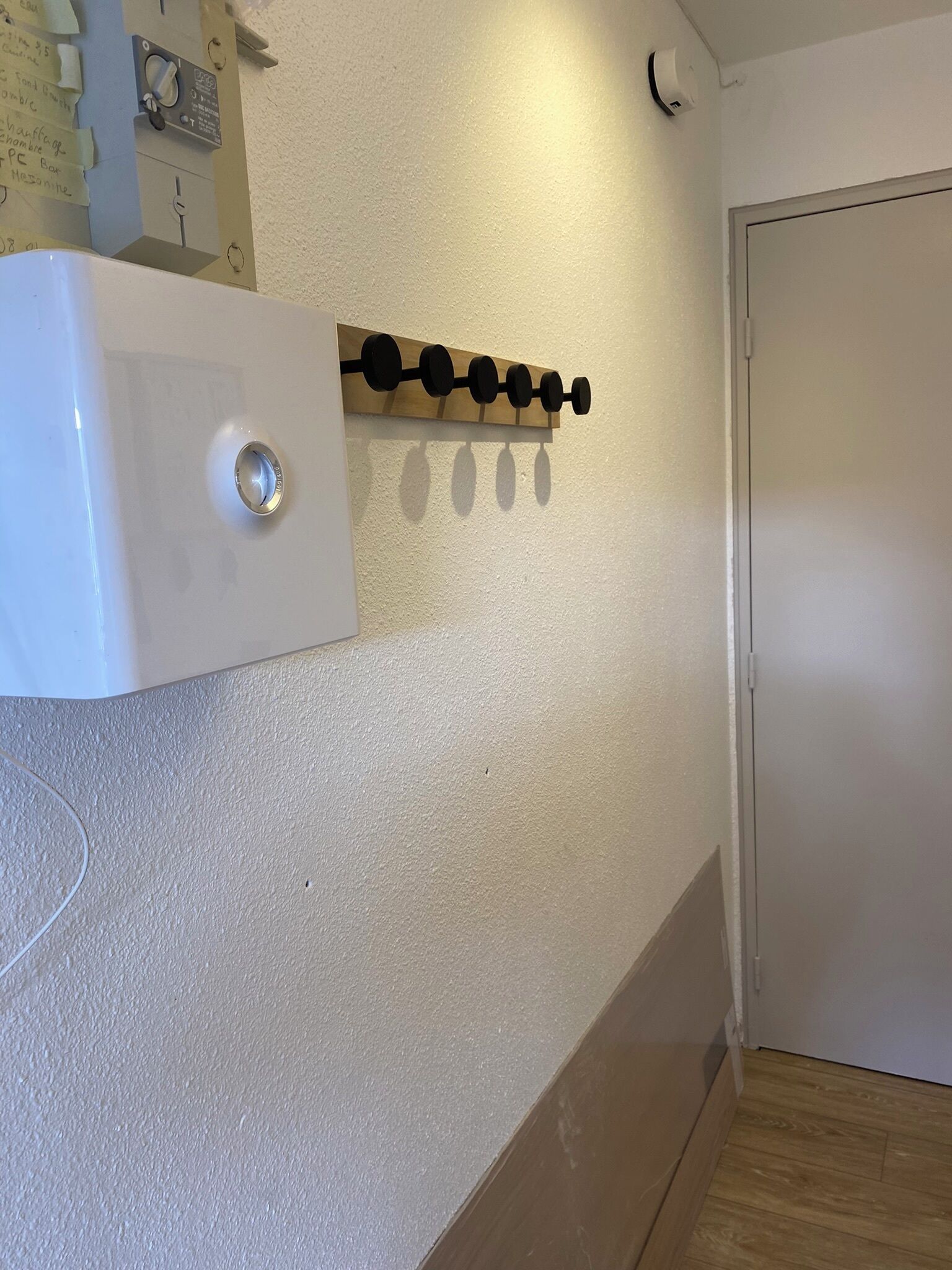 A white wall with a wooden shelf holding black circular objects, a white wall-mounted device, and a door.