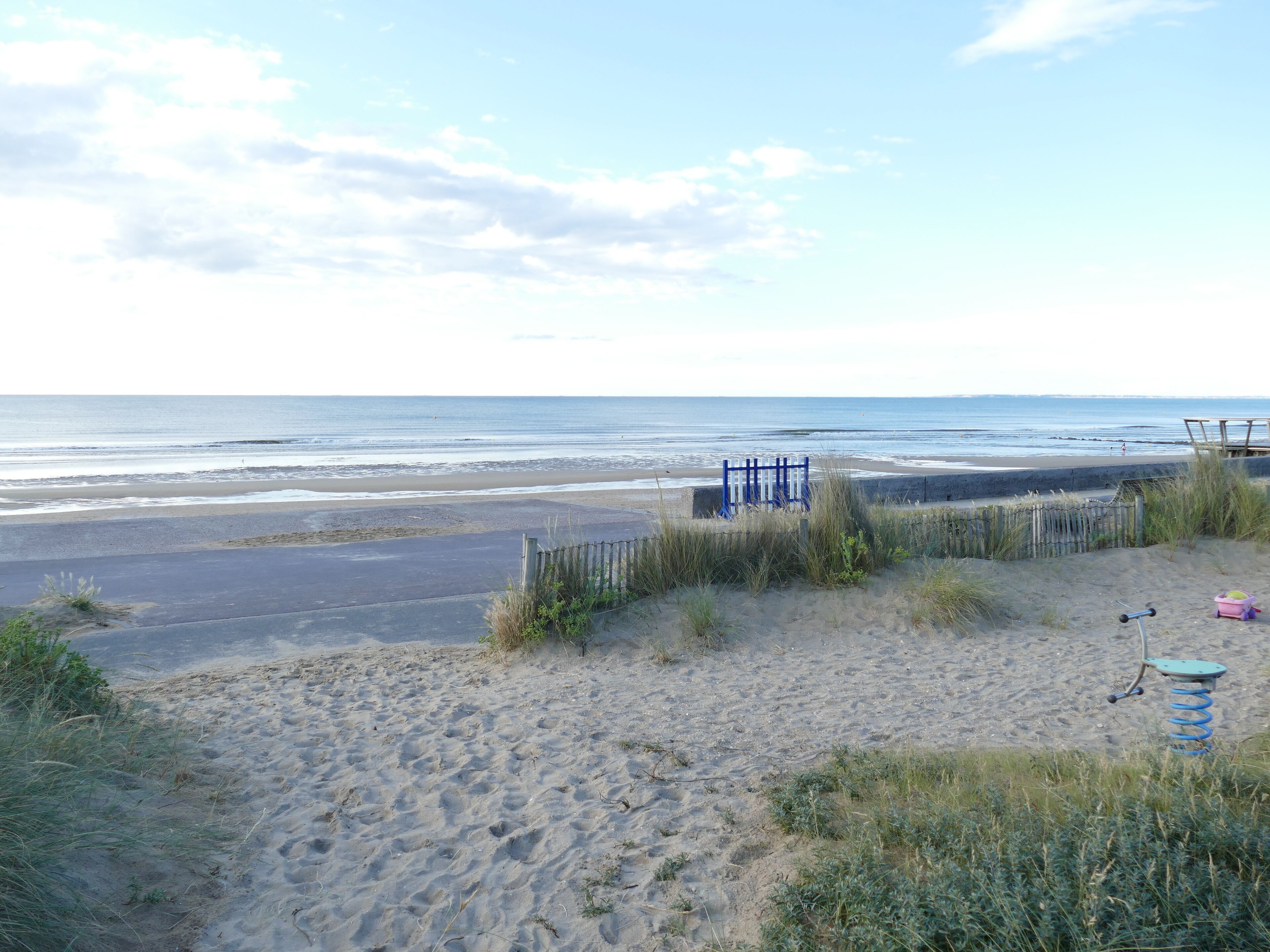 Une plage avec un sentier de sable menant à l’eau, une clôture bleue et une chaise de plage.