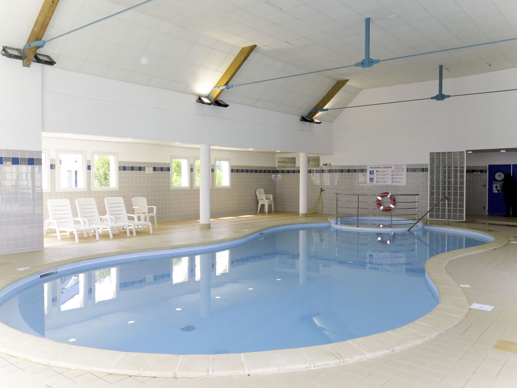 Une piscine intérieure avec un sol carrelé, une bouée de sauvetage et un panneau indiquant la zone de la piscine.