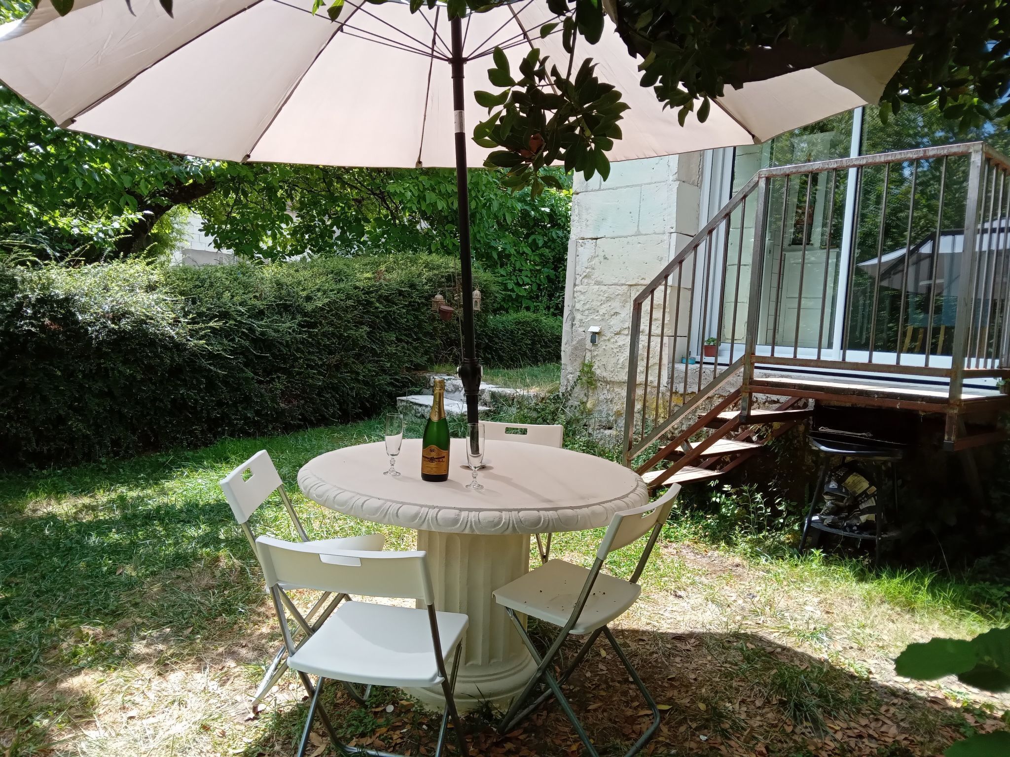 Eine Terrasse im Freien mit einem runden Tisch, zwei weißen Stühlen, einer Champagnerflasche und einem Sonnenschirm.