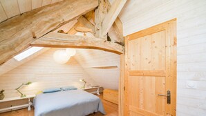 Ein gemütliches Schlafzimmer im Dachgeschoss mit schrägen Wänden, Holzbalken, einem Bett, einem Schreibtisch und einer Tür.