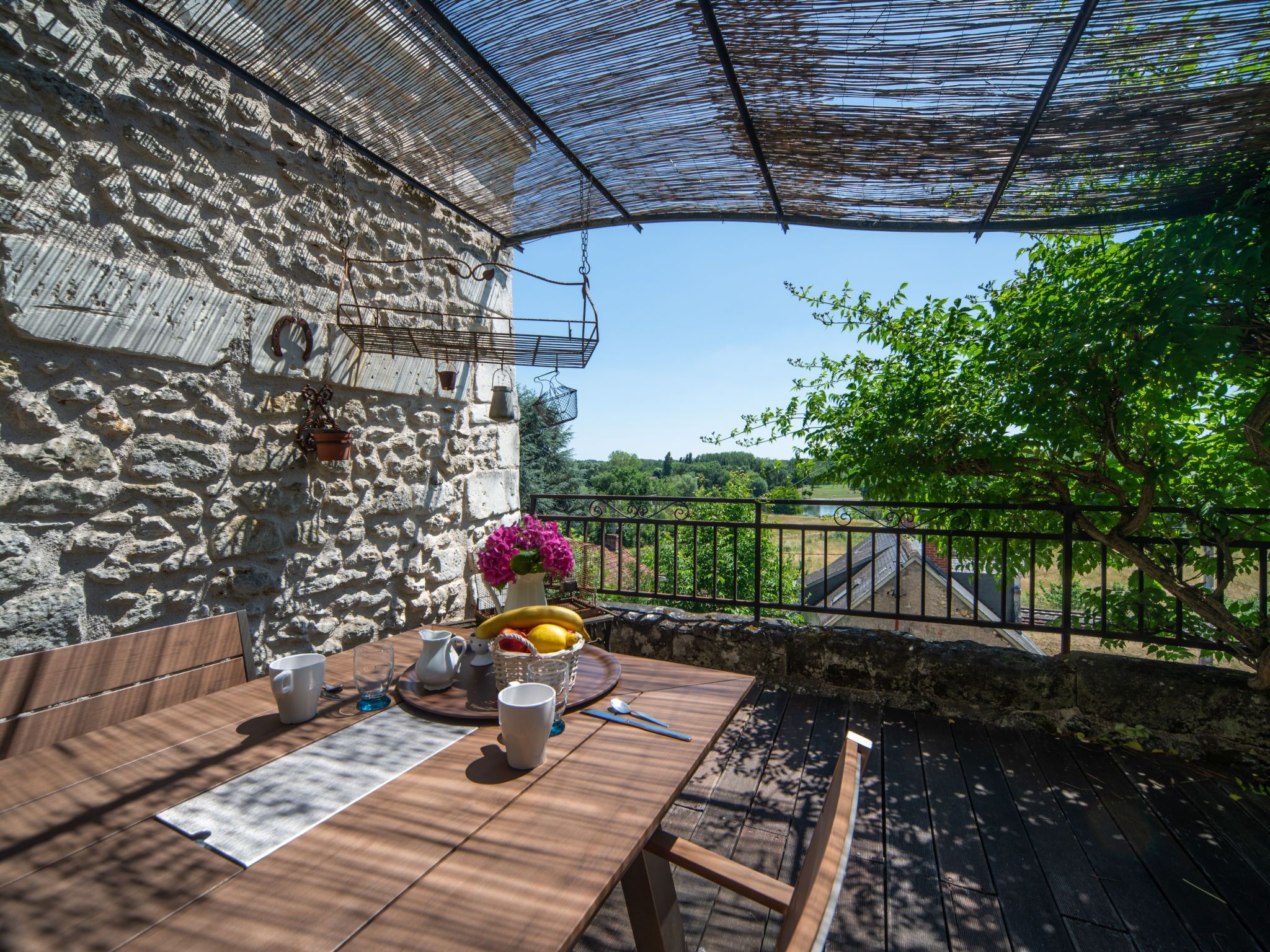 Un patio al aire libre con una mesa de madera dispuesta para comer, un muro de piedra y vistas a la vegetación y un puente.