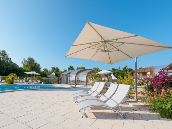 A swimming pool area with lounge chairs and umbrellas, surrounded by greenery and buildings. - Les Cottages du Lac M4 Privilège by Interhome (Parentis en Born)