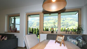 A room with a wooden dining table, a grey sofa, and a large window offering a view of a green landscape.