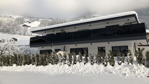 A modern two-story building with a flat roof, surrounded by snow-covered trees and a mountainous backdrop.