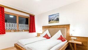 Un dormitorio con cama de madera, ropa de cama blanca, una ventana con cortinas rojas y un cuadro de montañas en la pared.