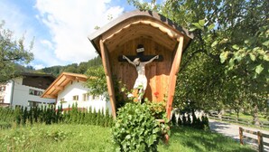 Una estructura de madera con una cruz y un cartel que dice 'INRI' en un área cubierta de césped, con una valla de madera y casas al fondo.