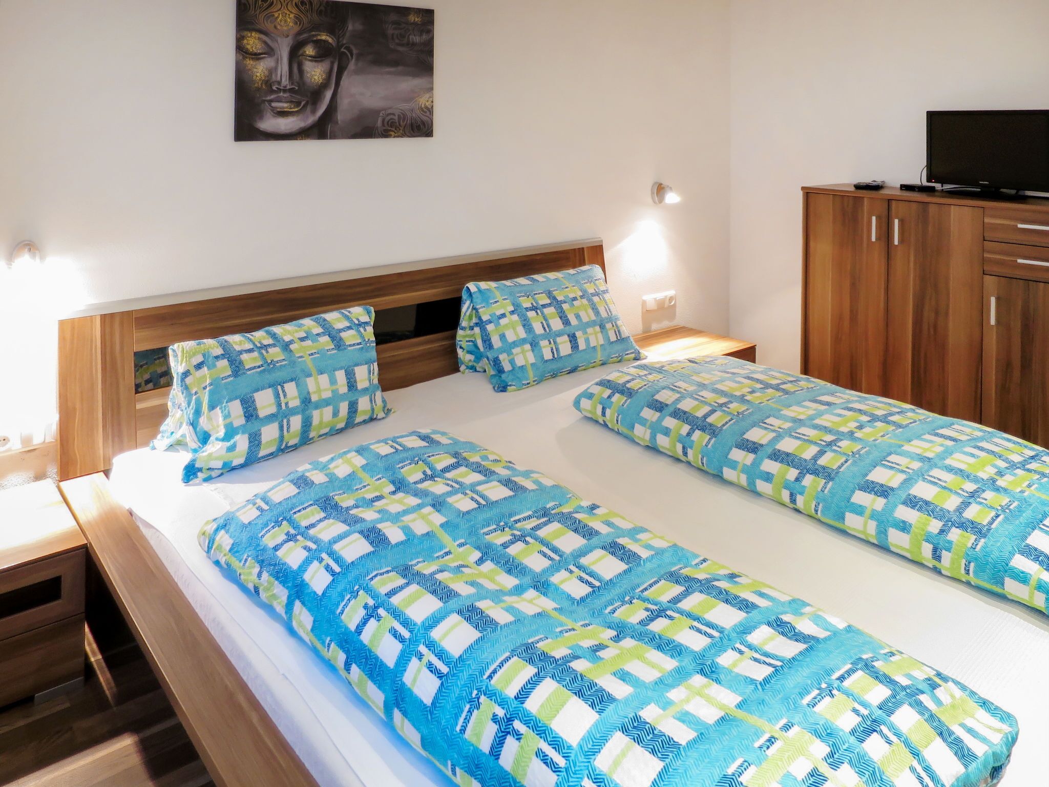 A double bed with blue and green patterned bedding, a wooden headboard, bedside tables with lamps, a wooden wardrobe, and a Buddha painting on the wall.