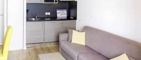 A modern living room with a grey sofa, a small white table, and a yellow chair. A kitchen with a microwave and a sink is visible in the background.