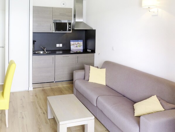 Una sala de estar moderna con un sofá gris, una mesita blanca y una silla amarilla. Se ve una cocina con un microondas y un fregadero al fondo.