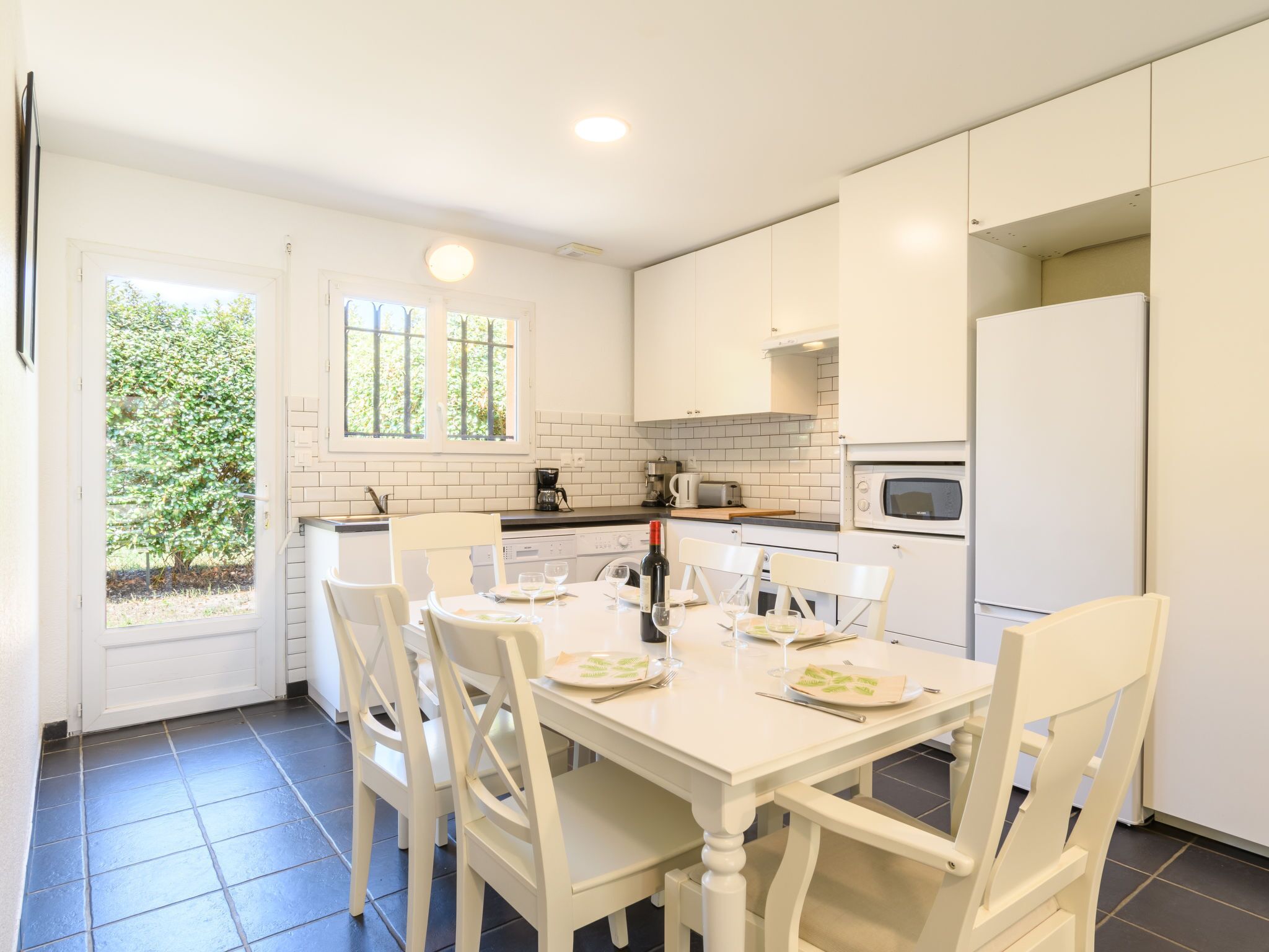 Una cocina moderna con armarios blancos, una mesa de comedor preparada para cuatro personas y un ventanal con vistas a zonas verdes.