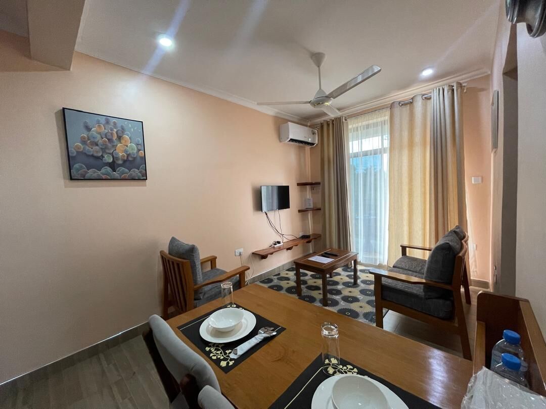 A dining area with a table set for two, a ceiling fan, a flat-screen TV, and a window with curtains.