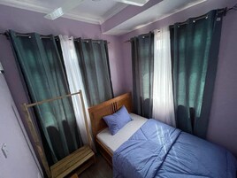 Una cama individual con cabecera de madera, una colcha azul y un almohadón en una habitación con paredes moradas y cortinas verdes.