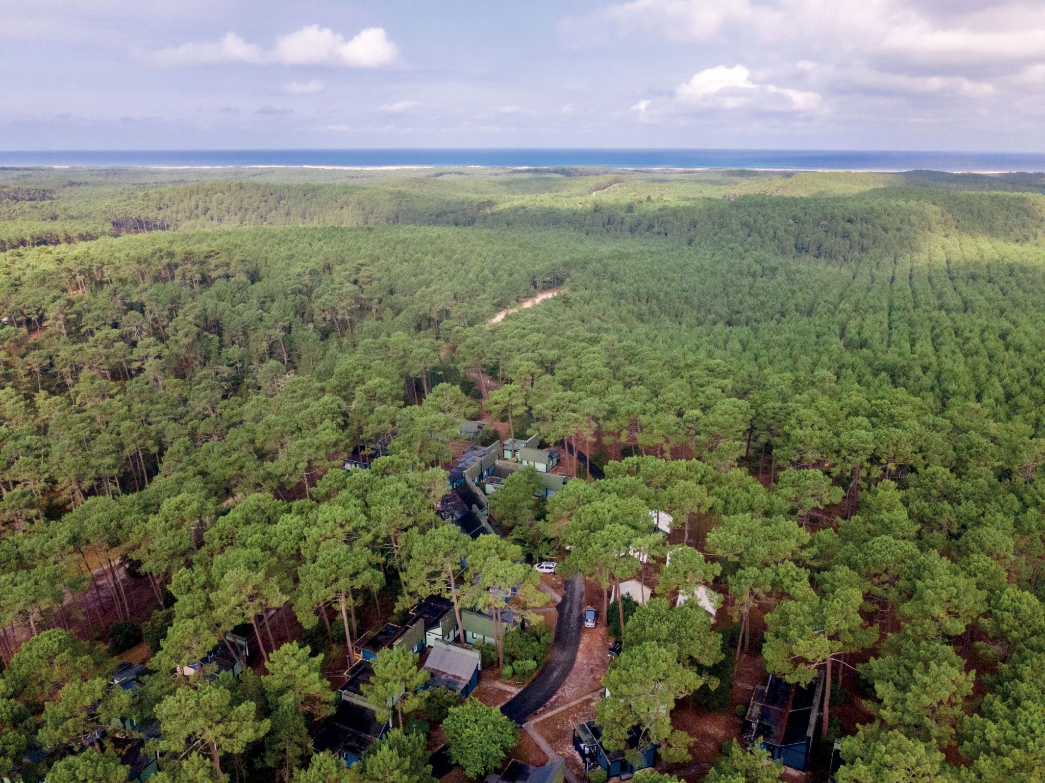 Une vue aérienne d’une zone boisée avec un petit groupe de bâtiments, probablement un complexe hôtelier ou un chalet, entouré d’arbres denses et sous un ciel dégagé.