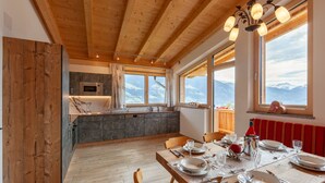Ett modernt kök med träbjälkar, en matplats med ett bord för fyra personer och stora fönster med utsikt över bergen.