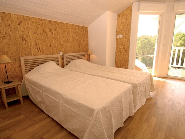 A room with two beds, wooden walls, a small table with a lamp, and a balcony with a view of trees.