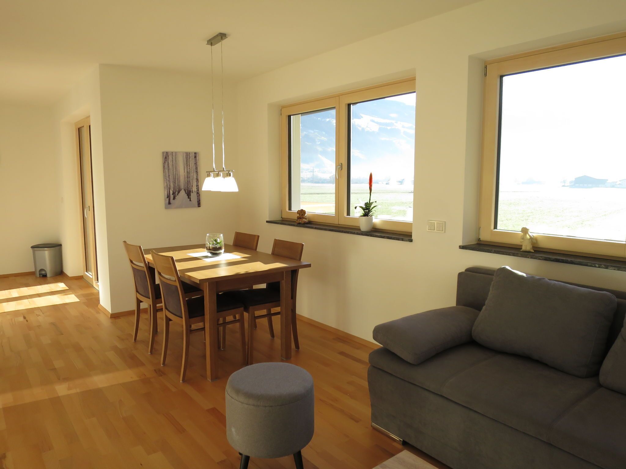 A spacious living area with a dining table, grey sofa, and large windows with a view of the outdoors.