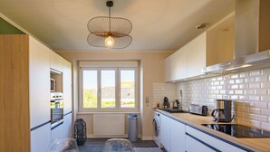 A modern kitchen with a large window, a hanging light fixture, a washing machine, and a coffee maker.
