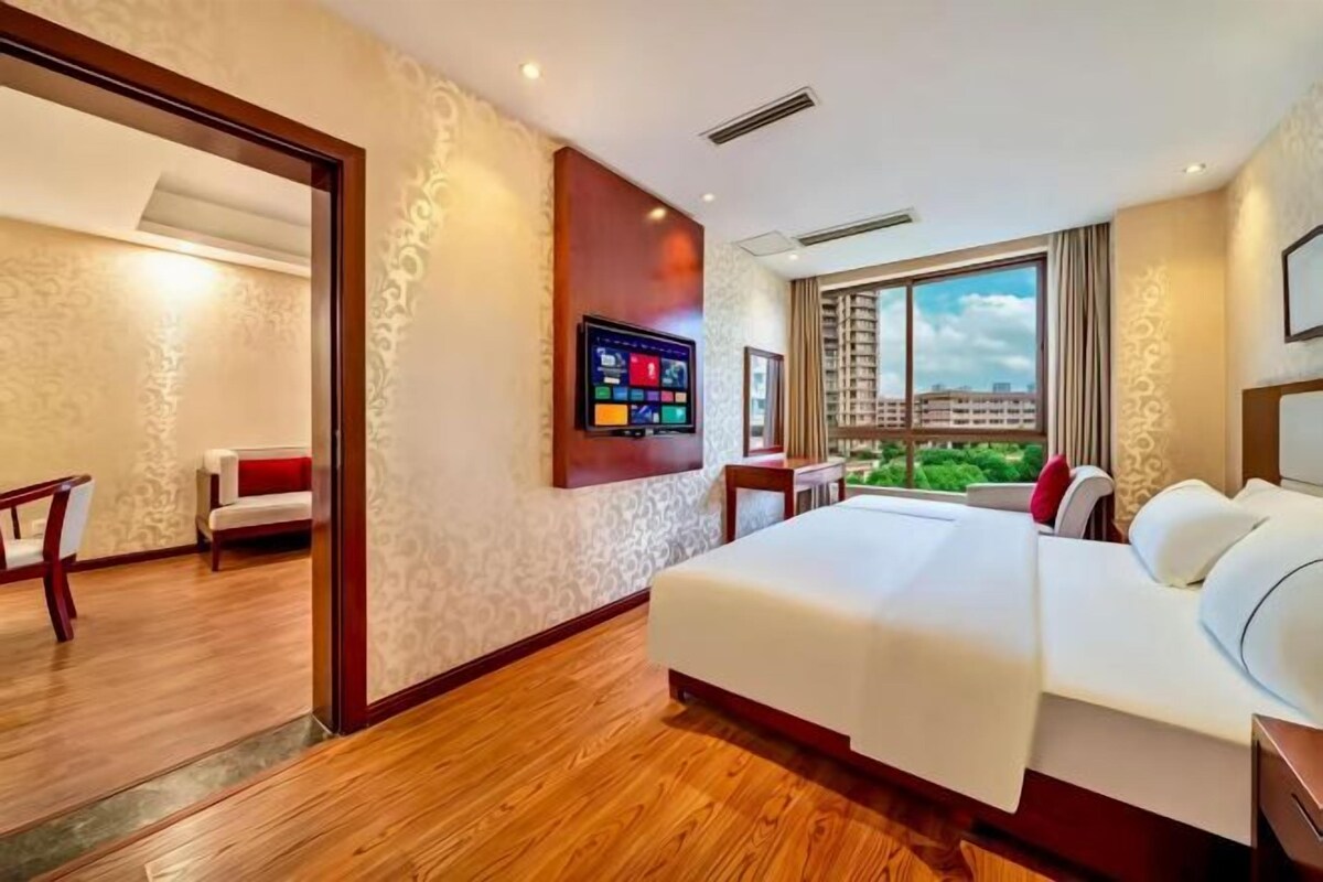 A hotel room with a large bed, a television mounted on the wall, and a window with a view of buildings.