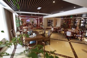 A spacious hotel dining area with multiple tables and chairs, a modern ceiling design, and a view of the adjacent room.