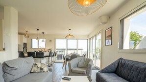 Un salon moderne avec des canapés gris, une table basse blanche sur laquelle repose un magazine, et un coin repas à l’arrière-plan.