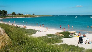 Une plage où l’on peut voir des gens se prélasser au soleil et nager, une eau d’un bleu limpide et des bateaux au loin.
