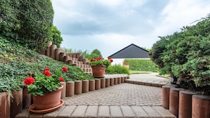 Ein gepflasterter Weg führt zu einem Gebäude mit dunklem Dach, umgeben von terrassierten Pflanzkästen mit roten Blumen und verschiedenen Sträuchern.