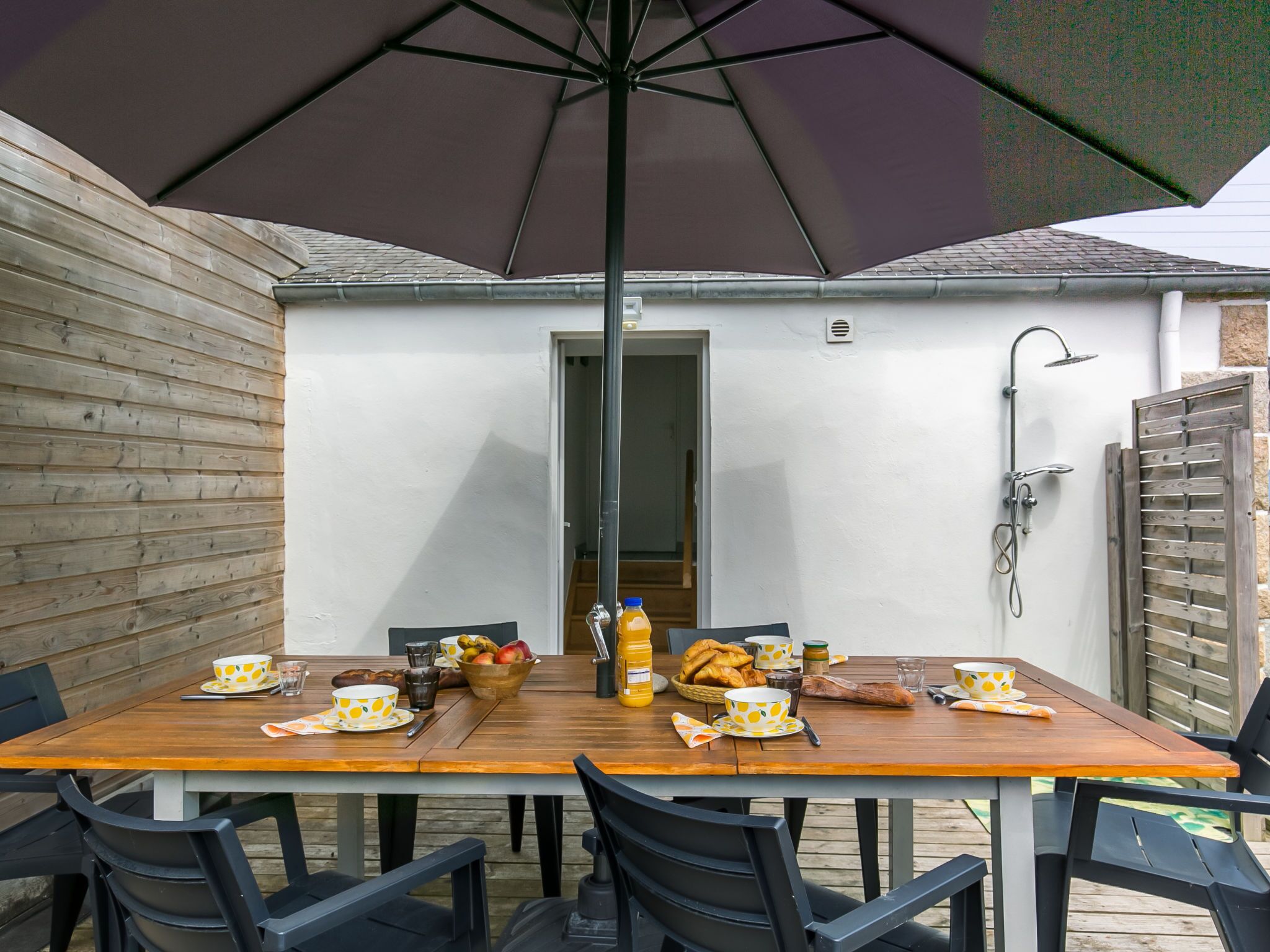 An outdoor dining area with a wooden table and chairs, a large umbrella, and a view of a white building with a wooden fence and a door.