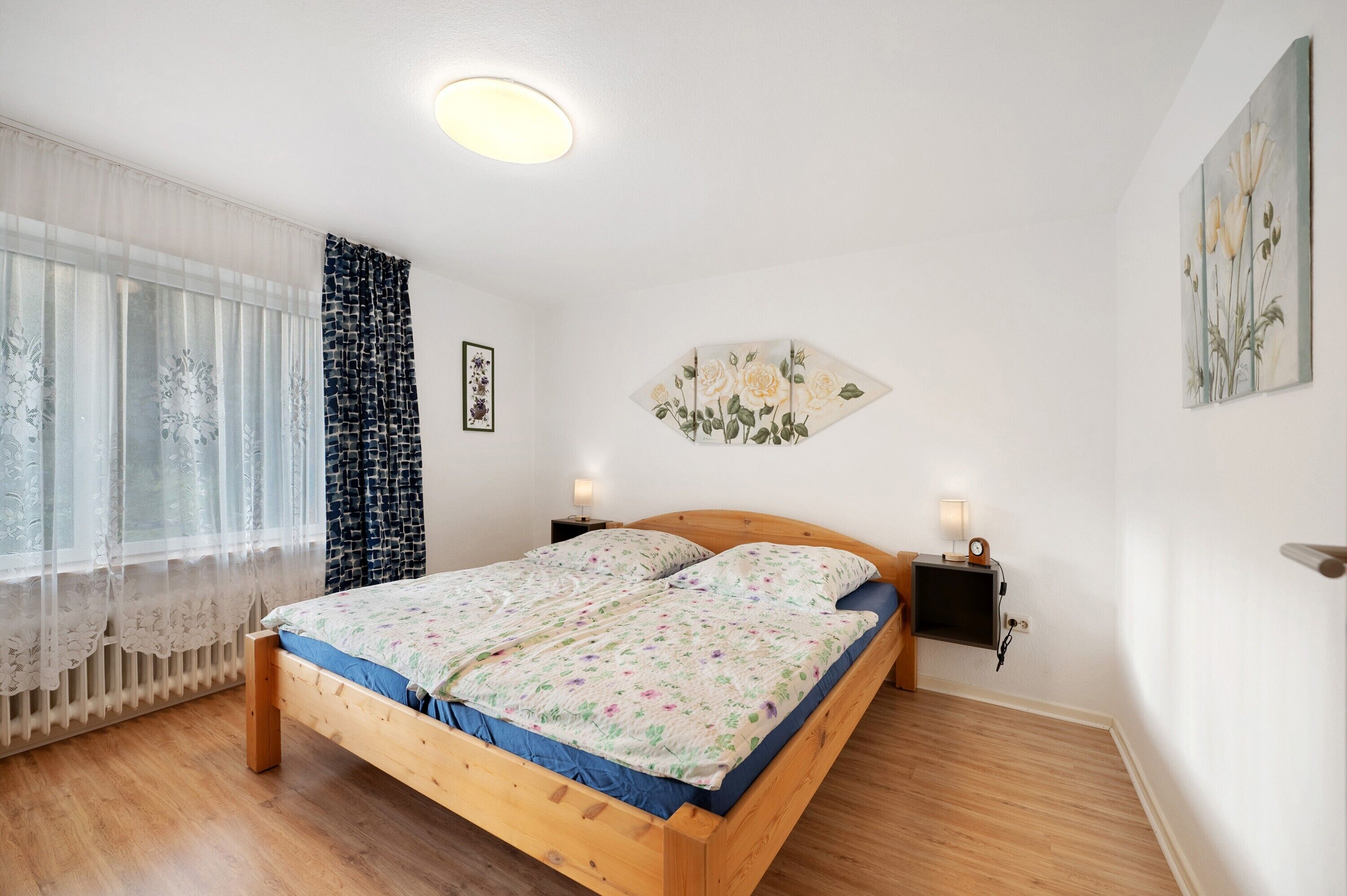 Un dormitorio con una cama de madera, dos mesitas de noche con lámparas, un televisor montado en la pared y una ventana con cortinas traslúcidas.