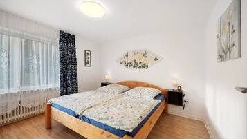 Un dormitorio con una cama de madera, dos mesitas de noche con lámparas, un televisor montado en la pared y una ventana con cortinas traslúcidas.