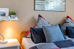 Habitación de hotel ordenada con cama, almohadas, lámpara de noche, planta pequeña y un cuadro en la pared.