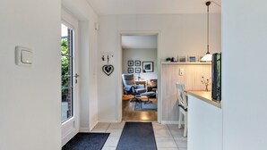 Un couloir mène à un espace de vie comprenant un canapé et des fauteuils, un coin cuisine avec un comptoir et un luminaire suspendu, ainsi qu’une porte ornée d’un motif en forme de cœur.