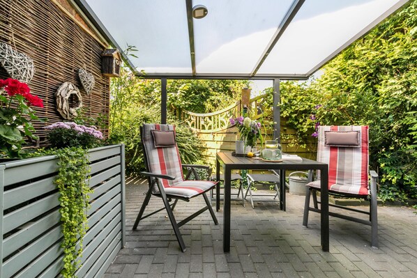 A covered patio with two chairs and a table, surrounded by greenery and a wooden fence.