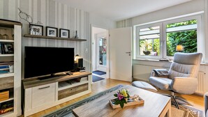 Un salon doté d’un téléviseur à écran plat, d’un meuble de rangement en bois, de bibliothèques, d’une table basse avec un vase, d’un fauteuil gris et d’une fenêtre donnant sur un paysage verdoyant.