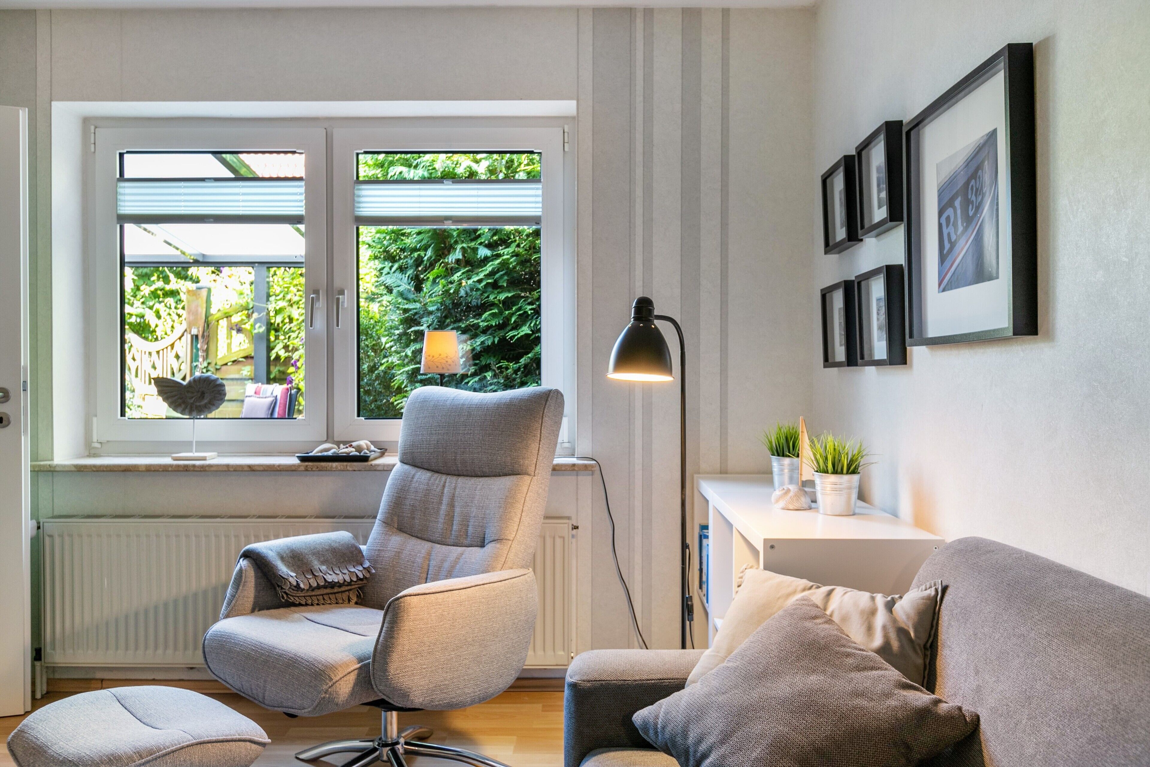 A modern living room with a grey armchair, a sofa, a standing lamp, and a window with a view of greenery.